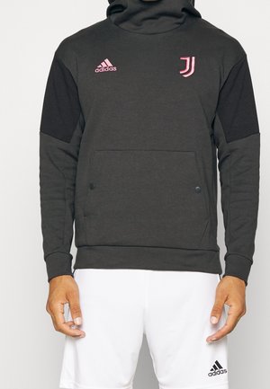 Mann trägt einen schwarzen Adidas-Hoodie mit Juventus-Logo und weiße Adidas-Shorts, steht vor einem schlichten Hintergrund.