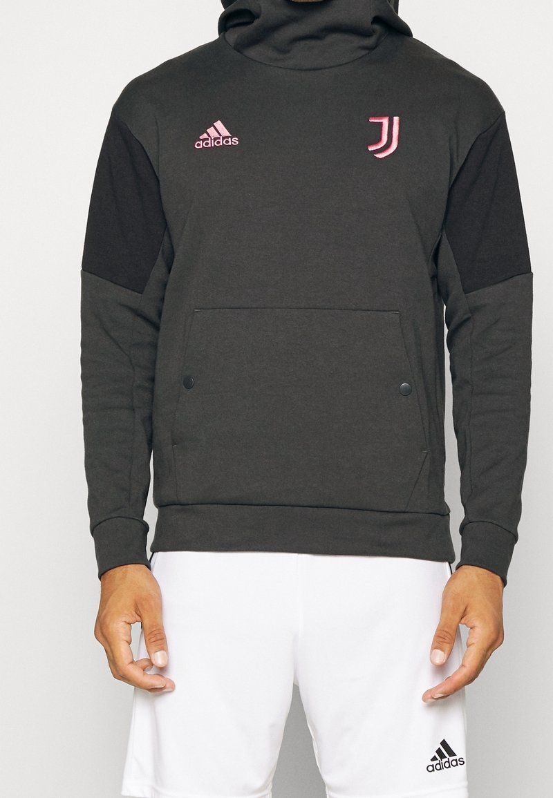 Homme portant un sweat à capuche noir Adidas avec le logo de la Juventus et un short blanc Adidas, debout sur un fond uni.