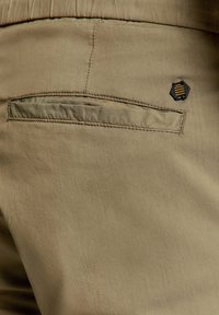 Khaki broek met een achterzak met een gelaagde flap, textuurstof en een zeshoekig zwart-gouden logoaccent nabij de zak.