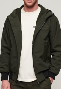 Olivengrøn hættetrøje med lynlås foran, elastiske manchetter og et diskret logo på brystet, lagdelt over en cremefarvet t-shirt.