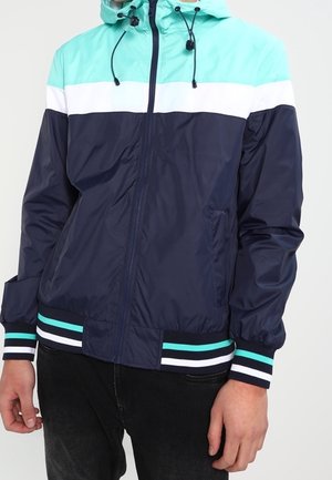 Uomo che indossa una giacca a vento color-block con sezioni verde menta, bianca e blu navy, polsini a righe e cappuccio con coulisse.