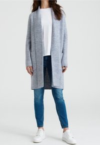 Cardigan lungo azzurro realizzato in morbido tessuto a maglia, con colletto a coste e due tasche frontali. Abbinato a una t-shirt bianca e jeans blu.