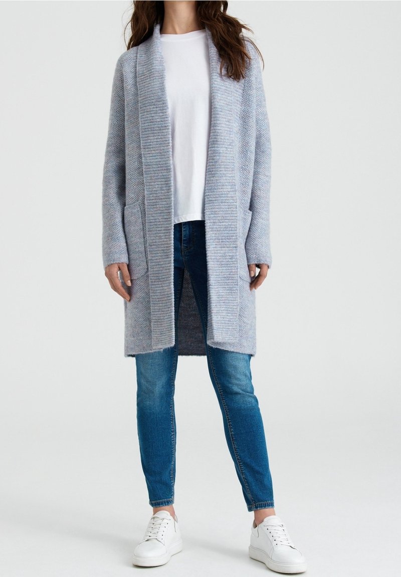 Cardigan lungo azzurro realizzato in morbido tessuto a maglia, con colletto a coste e due tasche frontali. Abbinato a una t-shirt bianca e jeans blu.