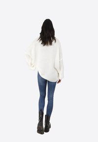 Maglione bianco oversize e strutturato con maniche ampie, abbinato a jeans blu attillati e stivali neri alla caviglia, visto da dietro.