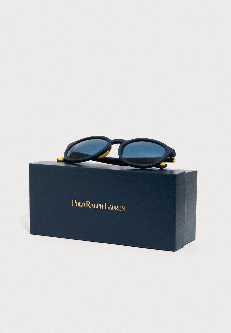 Black sunglasses with a rounded shape and blue lenses, displayed on a navy box featuring gold text. Yellow accents on the frame.