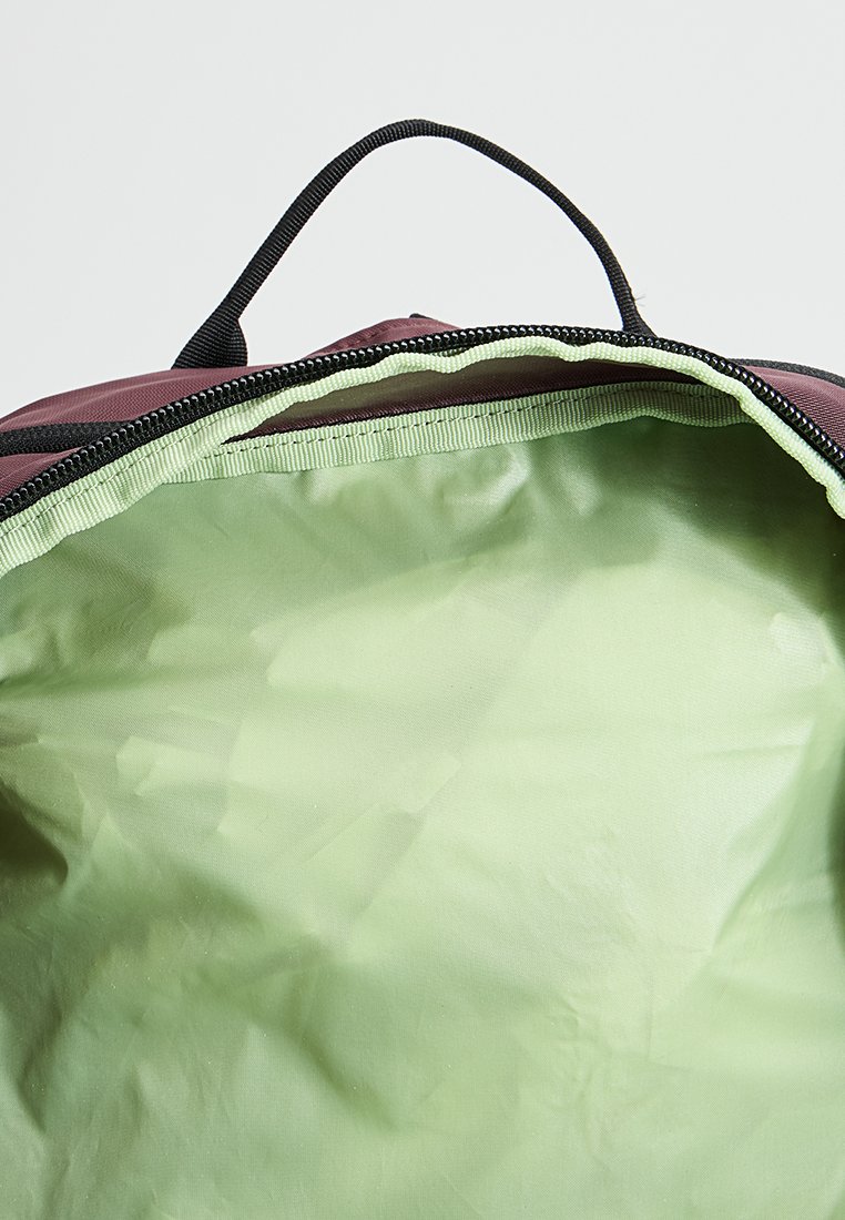 Mochila burdeos con cierre de cremallera, con un forro interior color verde claro que es suave y ligeramente brillante. Acentos de tela y cremallera en negro.