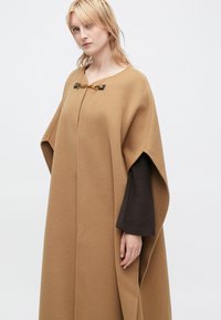 Καφέ μάλλινη κάπα με ρέουσα σχεδίαση, διαθέτει V-λαιμόκοψη, πλαϊνές σχισμές και διακοσμητική χρυσή αλυσίδα κούμπωμα στον γιακά.