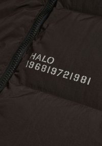 Zwarte pufferjack met een gladde, licht gekreukelde textuur. Voorzien van een ritsluiting en geprinte tekst "HALO 196819721981" in het grijs.