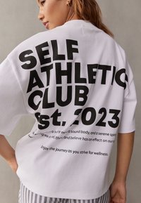 Vit oversized t-shirt med fet svart text på baksidan, inklusive "SELF ATHLETIC CLUB" och "est. 2023." Mjuk tyg, avslappnad passform.
