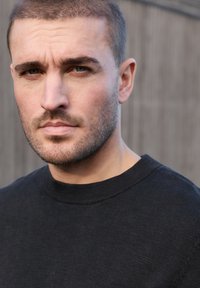 Gros plan sur un homme sérieux aux cheveux courts et à la légère barbe de quelques jours, portant un pull noir à col rond, sur un fond neutre.