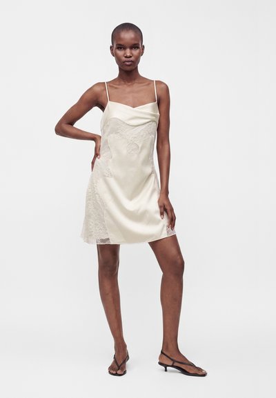 Femme portant une robe nuisette en satin crème sans manches avec des détails en dentelle et des sandales noires à talons bas, debout, une main sur la hanche.