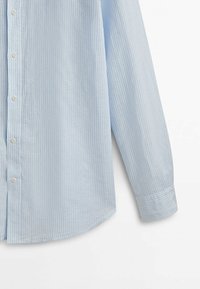 Chemise à manches longues bleu clair avec fines rayures verticales blanches, boutons visibles et manchette retroussée sur un fond blanc.
