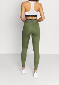 Olivově zelené legíny s hladkou texturou, vysokým pasem a bočními kapsami. V kombinaci s bílou sportovní podprsenkou s černým pruhem.