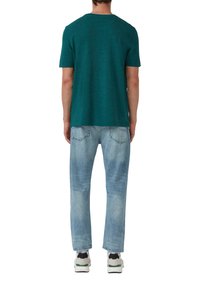 Turkusowy t-shirt z krótkim rękawem i okrągłym dekoltem, w zestawieniu z jasnoniebieskimi, lekko przetartymi dżinsami. Model stoi tyłem, mając na sobie białe trampki.