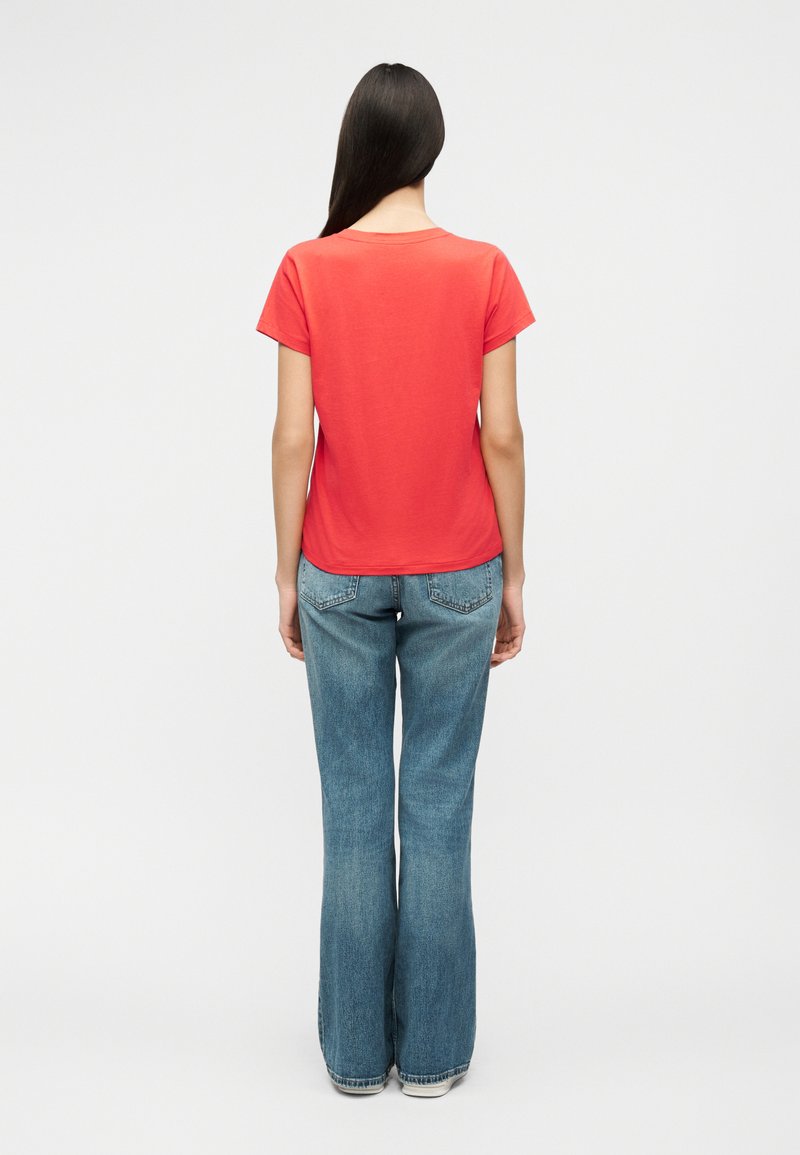 Vrouw met lang donker haar, gekleed in een effen rood T-shirt met korte mouwen en blauwe wijde jeans, staande met de rug naar voren op een witte achtergrond.