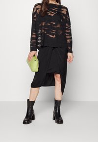 Svart genomskinlig långärmad topp med utskurna mönster, tillsammans med en svart knuten kjol. Håller en grön imitationsläderväska och bär svarta klackboots.