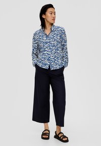 Langärmliges Hemd mit einem blau-weißen abstrakten Muster, aus leichtem Stoff, mit knöpfbarem Kragen, kombiniert mit marineblauen weit geschnittenen Hosen und schwarzen Sandalen.