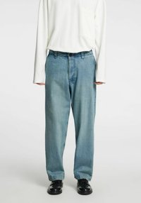 Svetlo modre kavbojke z ohlapnim krojem, z zadrgo na gumbe, z zankami za pas in rahlo zavihanimi robovi, nošene s črnimi čevlji.