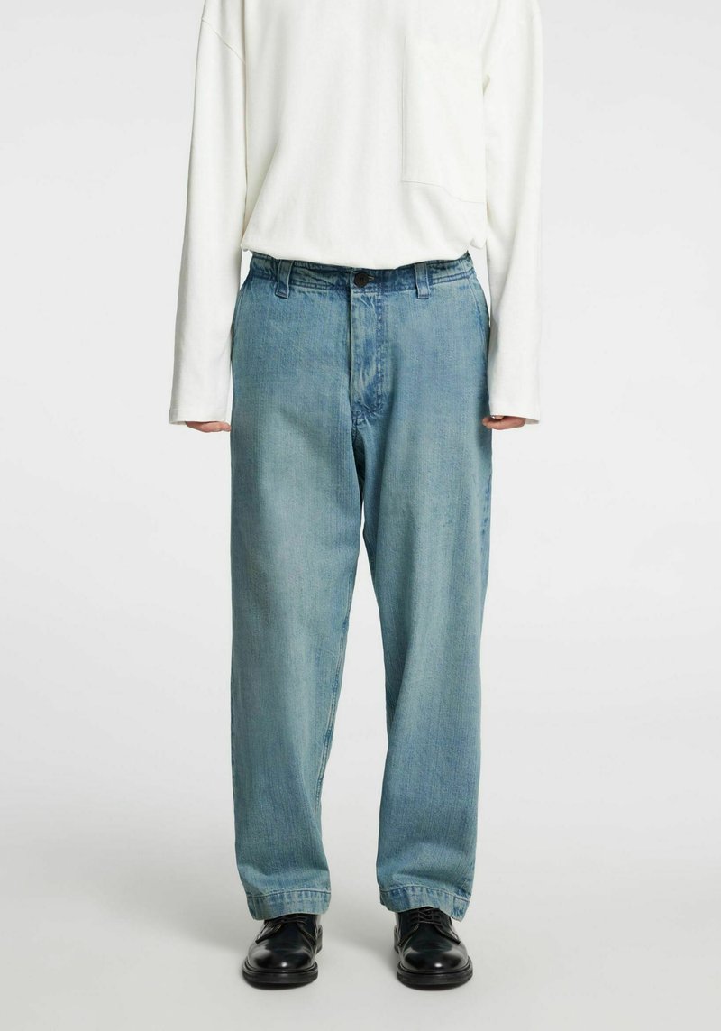 Svetlo modre kavbojke z ohlapnim krojem, z zadrgo na gumbe, z zankami za pas in rahlo zavihanimi robovi, nošene s črnimi čevlji.