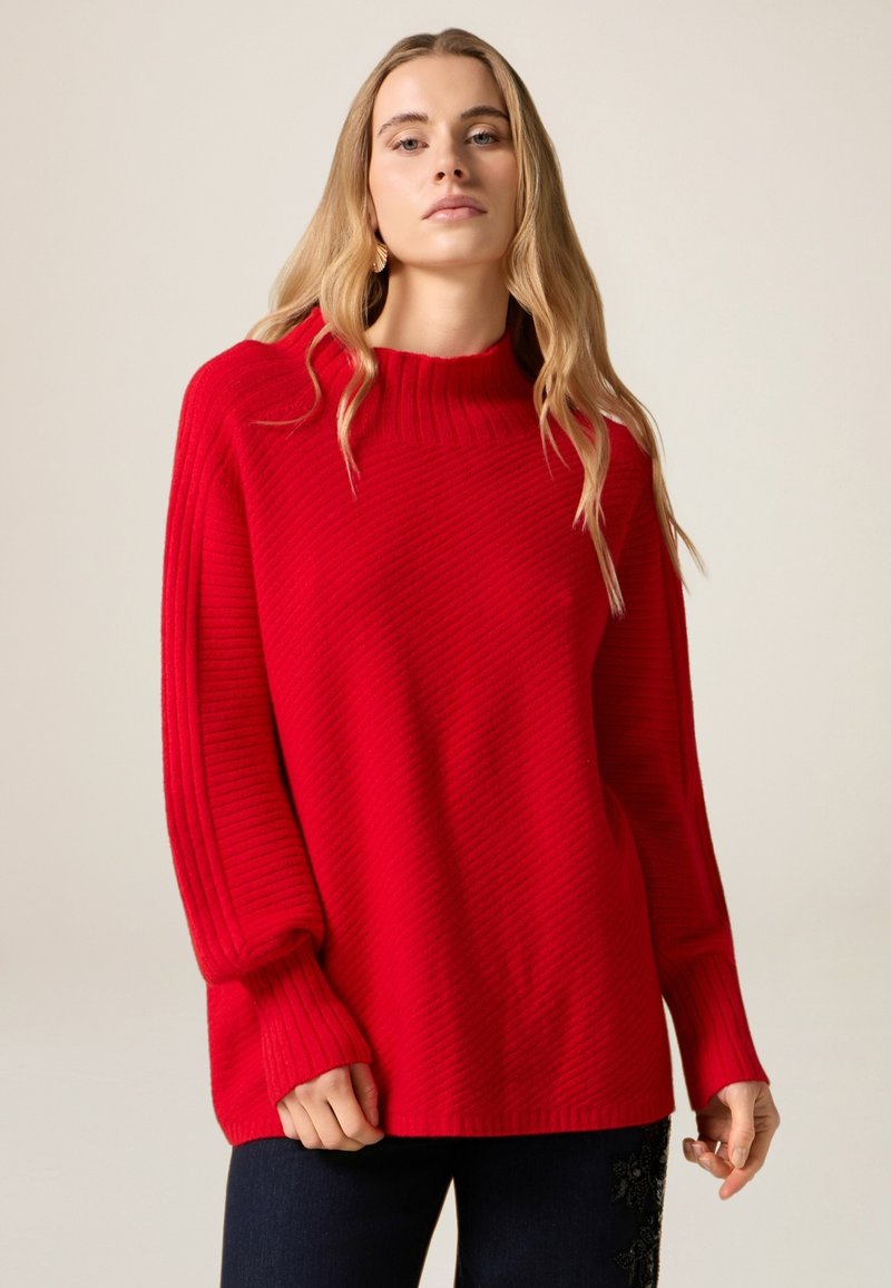 Maglione rosso in maglia a coste con collo alto e vestibilità ampia, caratterizzato da maniche lunghe e tessuto strutturato. Indossato con jeans scuri.
