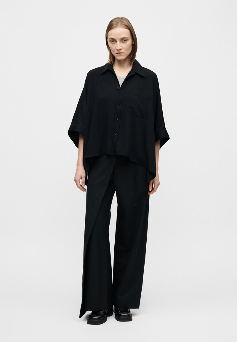 Chemise noire oversize à boutons avec manches courtes, associée à un pantalon noir à jambes larges avec une fente latérale, en tissu lisse.