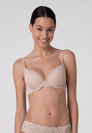 Donna con capelli scuri raccolti, che indossa un reggiseno e slip in pizzo beige coordinati, sorridente su uno sfondo chiaro e uniforme.