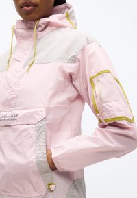 Άτομο που φοράει μπουφάν Columbia Sportswear σε παστέλ αποχρώσεις ροζ και μπεζ με τσέπη από δίχτυ στο μανίκι, με το χέρι στην εμπρόσθια τσέπη, στέκεται μπροστά σε λευκό φόντο.