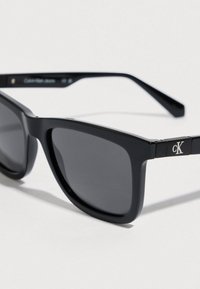 Mustat nelikulmaiset aurinkolasit tummilla linsseillä, joissa on muovikehykset ja hillitty hopeinen logo laajassa osassa. Tyylikäs, moderni muotoilu.