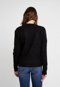 Woman with brown hair wearing gold hoop earrings, a black long-sleeve sweater, and blue jeans, facing away against a plain background.