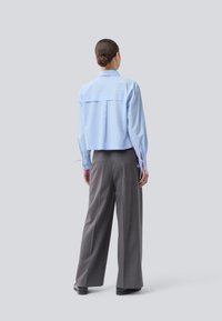 Frau mit einem Dutt, die ein hellblaues Langarmshirt und weite graue Hosen trägt, steht mit dem Rücken zu einem schlichten hellen Hintergrund.