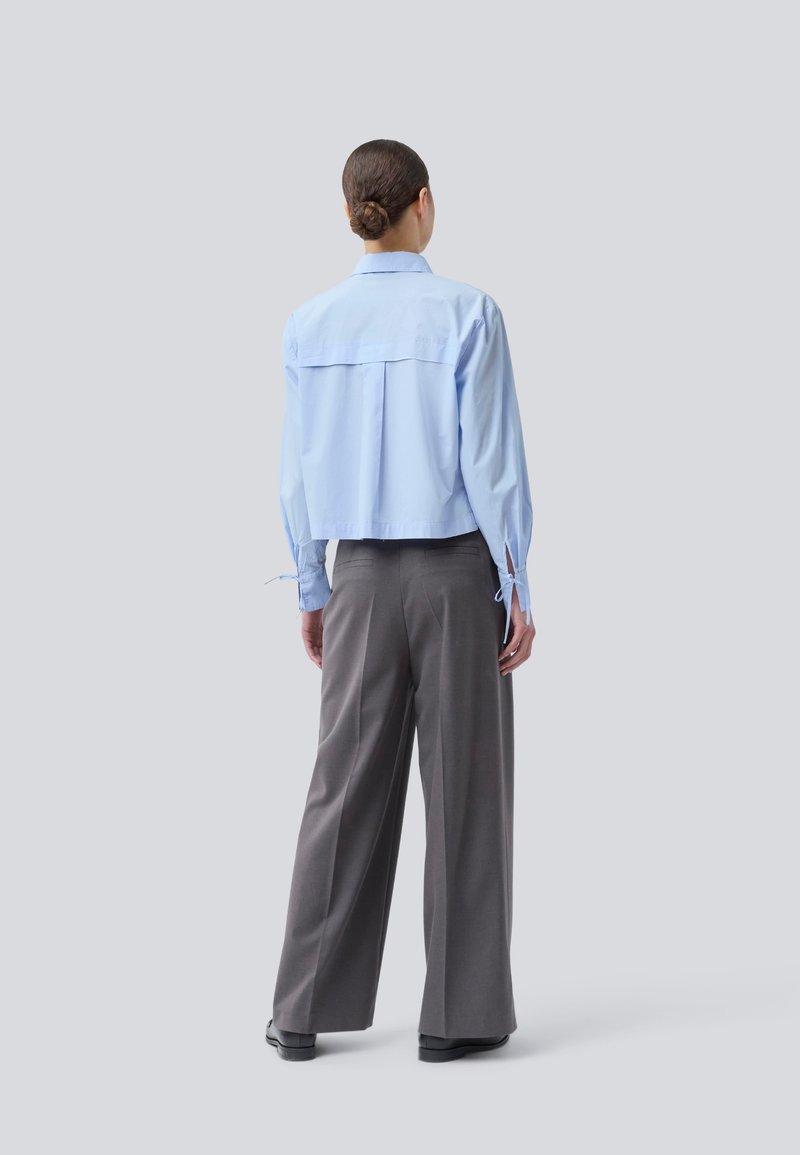 Frau mit einem Dutt, die ein hellblaues Langarmshirt und weite graue Hosen trägt, steht mit dem Rücken zu einem schlichten hellen Hintergrund.