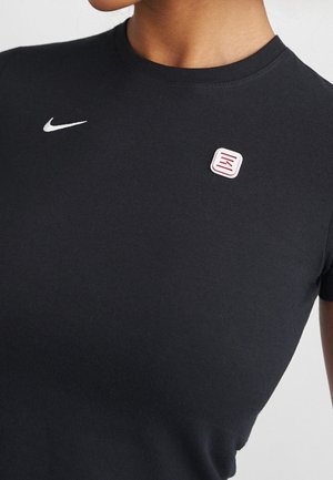 Osoba nosząca czarną koszulkę Nike z małym białym logo typu swoosh na lewej piersi oraz czerwono-białą przypinką powyżej.