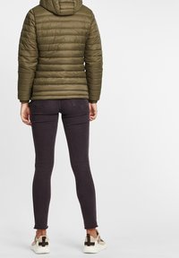 Doudoune vert olive avec capuche, associée à un jean skinny violet foncé et des baskets beiges avec un accent noir au talon.