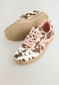Ein Paar low-top Sneakers mit braunem und weißem Kuhfellmuster, pinken Schnürsenkeln, pinken Seitenstreifen und beigen strukturierten Gummisohlen auf einem hellen Hintergrund.