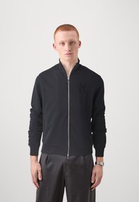 Juodas zip-up džemperis su stovinčiu apykakle, pagamintas iš minkštos medžiagos. Savybės: subtilus logotipas krūtinėje ir raišteliai rankogaliuose.