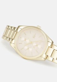 Reloj dorado con una pulsera metálica lisa. La esfera redonda presenta una carátula clara, tres subesferas y marcadores de hora que parecen diamantes. Bisel texturizado.