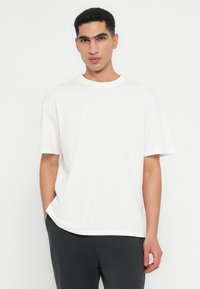 Νεαρός άνδρας με κοντά σγουρά μαλλιά, φορώντας ένα απλό άσπρο υπερμεγέθες μπλουζάκι και μαύρο παντελόνι, σε άσπρο φόντο.