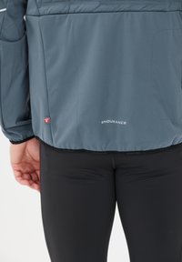Nærbillede af en person iført en grå jakke med "ENDURANCE" logo og sorte træningsbukser, med hånden hvilede ved siden af på en ensfarvet baggrund.