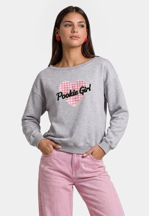 Jonge vrouw met hartvormige oorbellen, een grijze trui met een roze geruit hart en de tekst "Pookie Girl", en een roze broek met handen in de zakken.
