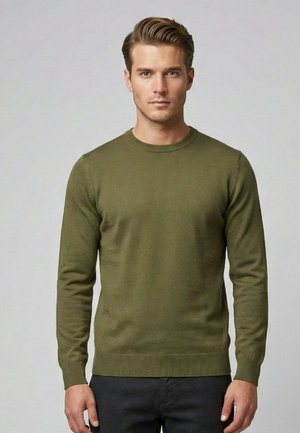 Uomo con capelli corti e acconciati, che indossa un maglione verde oliva a maniche lunghe e pantaloni neri, in piedi contro uno sfondo grigio uniforme.