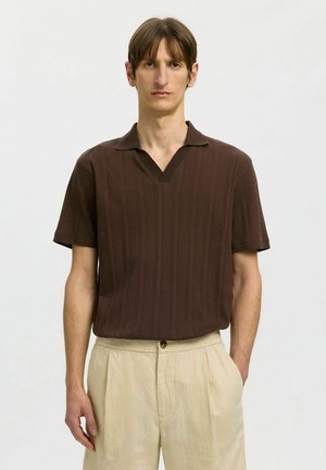 Homme portant une chemise marron à manches courtes avec col, rentrée dans un pantalon beige taille haute, sur un fond clair uni.