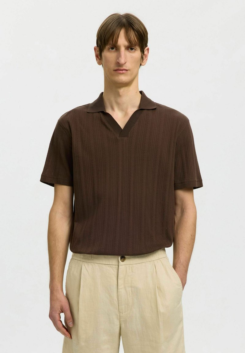 Homme portant une chemise marron à manches courtes avec col, rentrée dans un pantalon beige taille haute, sur un fond clair uni.