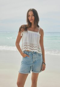 Witte mouwloze top met kantafwerking en gelaagd ontwerp, gecombineerd met lichtblauwe high-waisted denim shorts. Oceaan op de achtergrond.