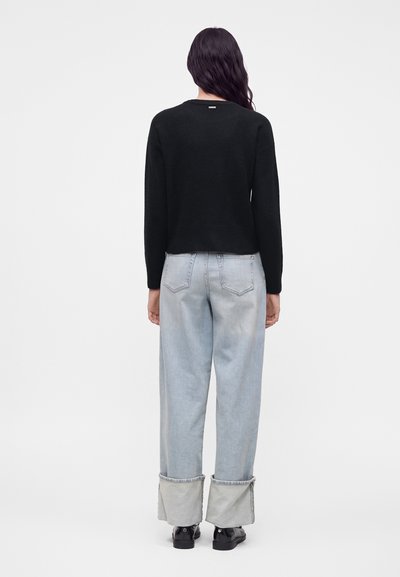 Personne aux longs cheveux foncés portant un pull noir, un jean large bleu clair avec des revers, et des chaussures noires, debout de dos sur un fond uni.