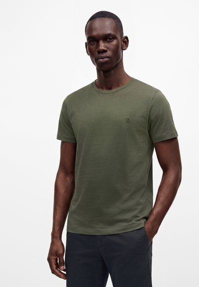 Homme noir portant un t-shirt vert olive à manches courtes et un pantalon foncé, debout avec une main dans la poche sur un fond clair uni.