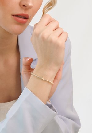 Gold bracelet with a row of small, clear gemstones on a delicate chain, worn on a wrist with light skin and a pale blue blouse.