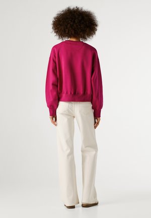 Persoon met krullend haar in een fuchsia lange mouwen sweatshirt en witte hoge taille brede broek, staand met de rug naar de camera tegen een witte achtergrond.