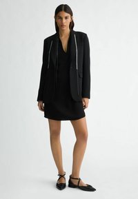 Crni blejzer s ukrasnim srebrnim detaljima, nosi se preko crne haljine. Model nosi crne ravne cipele s mašnom. Glatka tekstura.
