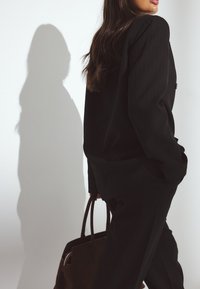 Kobieta w czarnym garniturze w prążki, trzymająca brązową skórzaną torebkę, stojąca bokiem z cieniem rzucanym na białą ścianę.