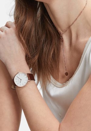 Femme portant un haut blanc sans manches, une montre à bracelet en cuir marron au poignet, et un collier en chaîne en or rose avec un pendentif circulaire.